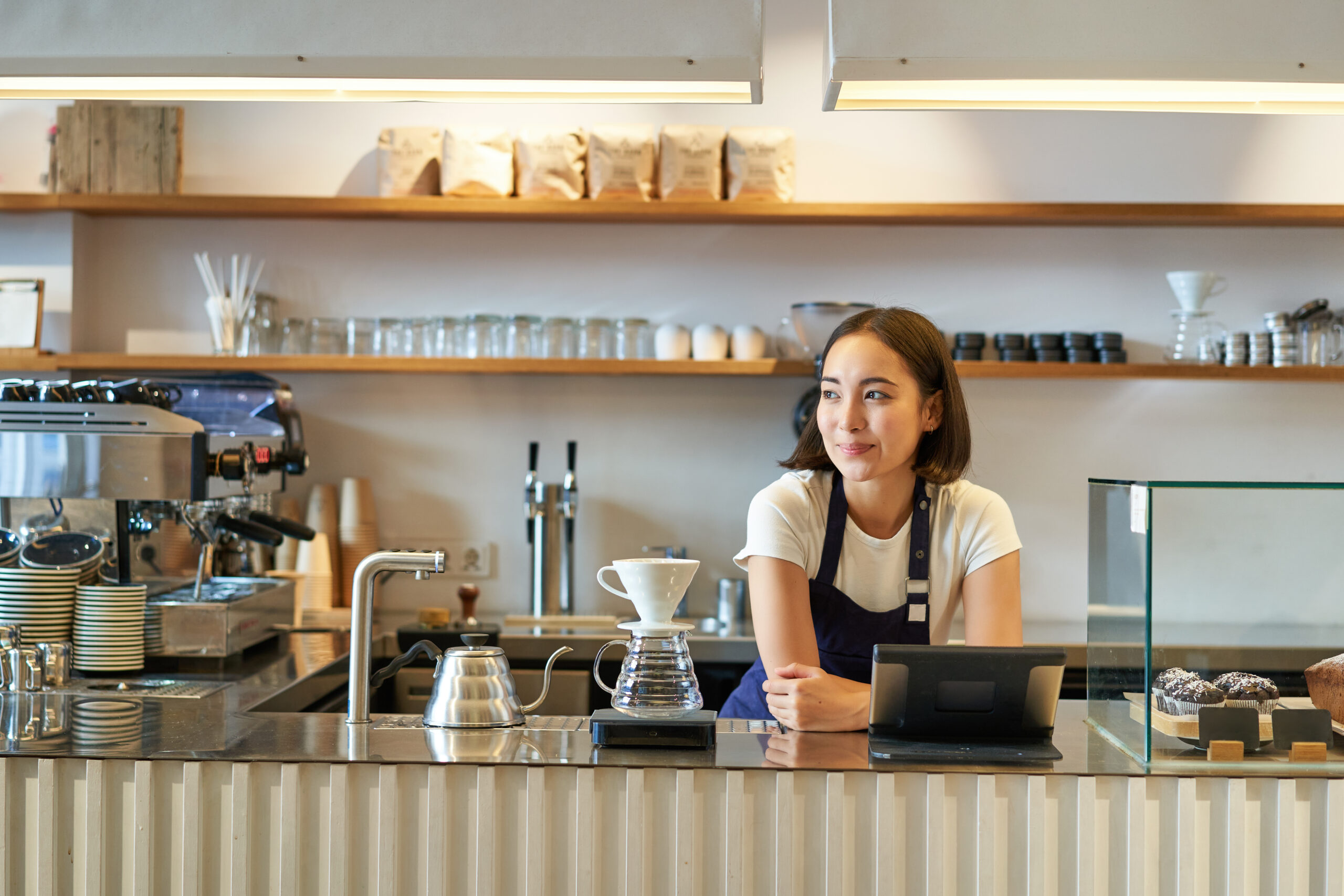 Happy smiling asian barista, girl behind counter, working with POS terminal and brewing filter kit, making coffee in cafe